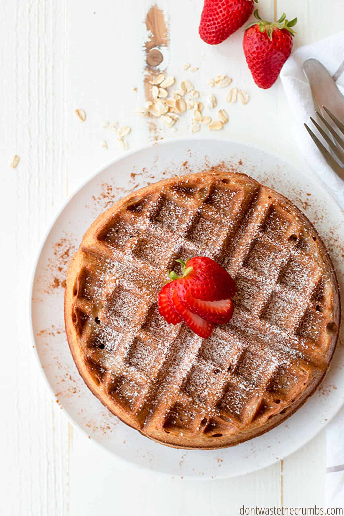A freshly toasted cinnamon oatmeal waffle, golden brown, and dusted with ground cinnamon and powdered sugar. Fresh strawberries sit atop the waffles.