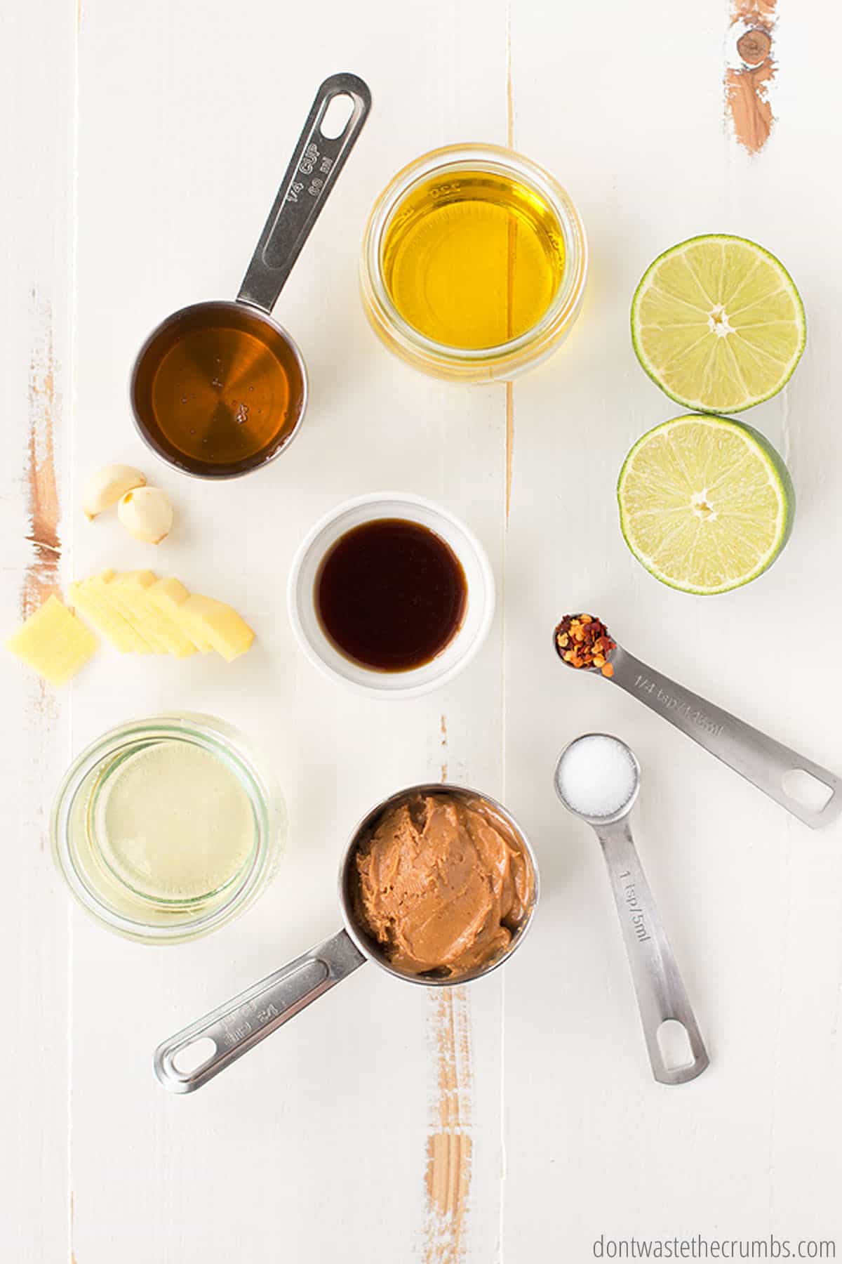 Shown are all the measured ingredients needed for Thai peanut sauce: halved limes, honey, rice vinegar, soy sauce, garlic, sliced ginger, red pepper, salt, peanut butter, sugar, and maple syrup.