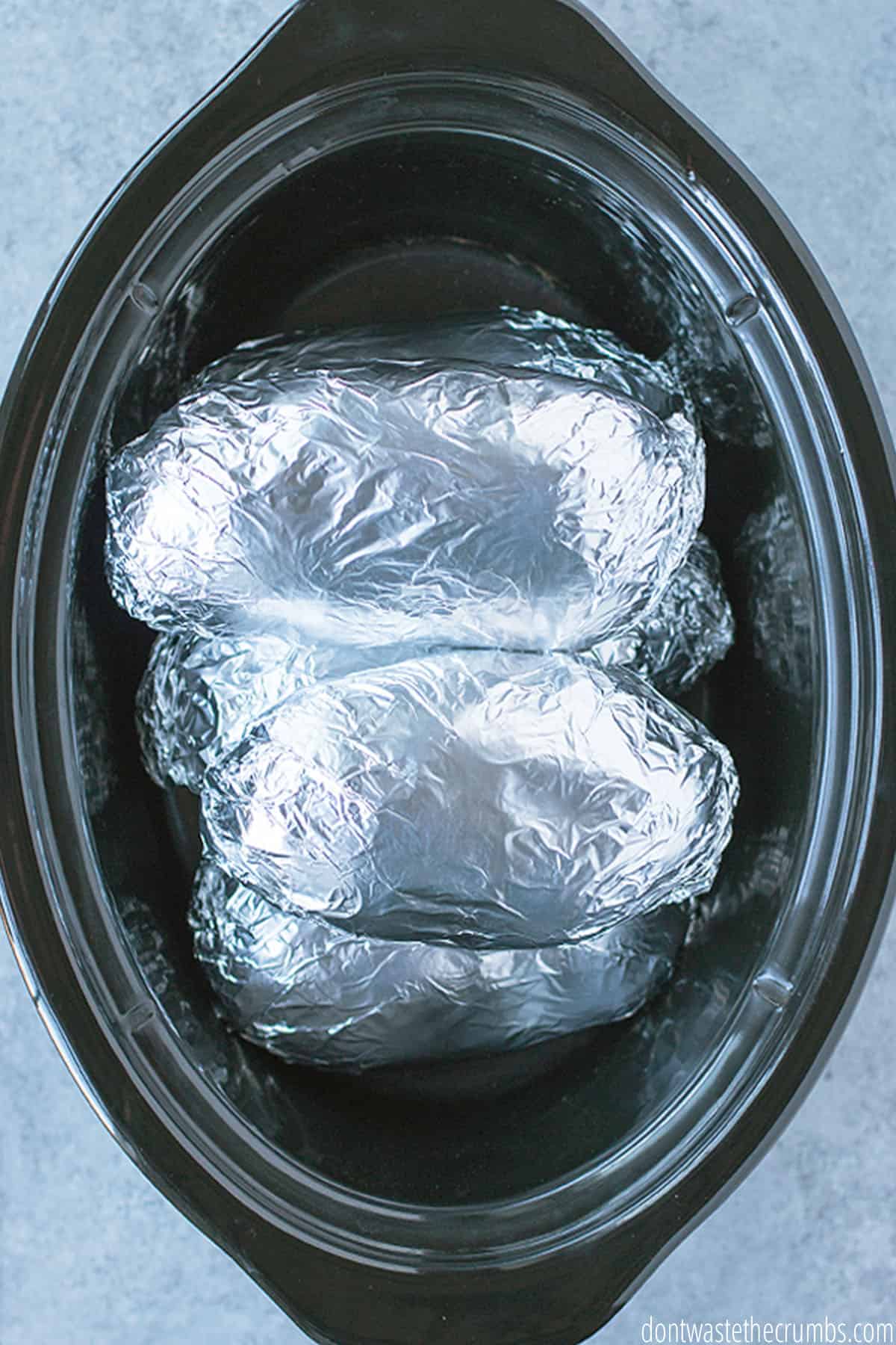 A black oval slow cooker holds 6 potatoes wrapped in foil ready for slow cooking.