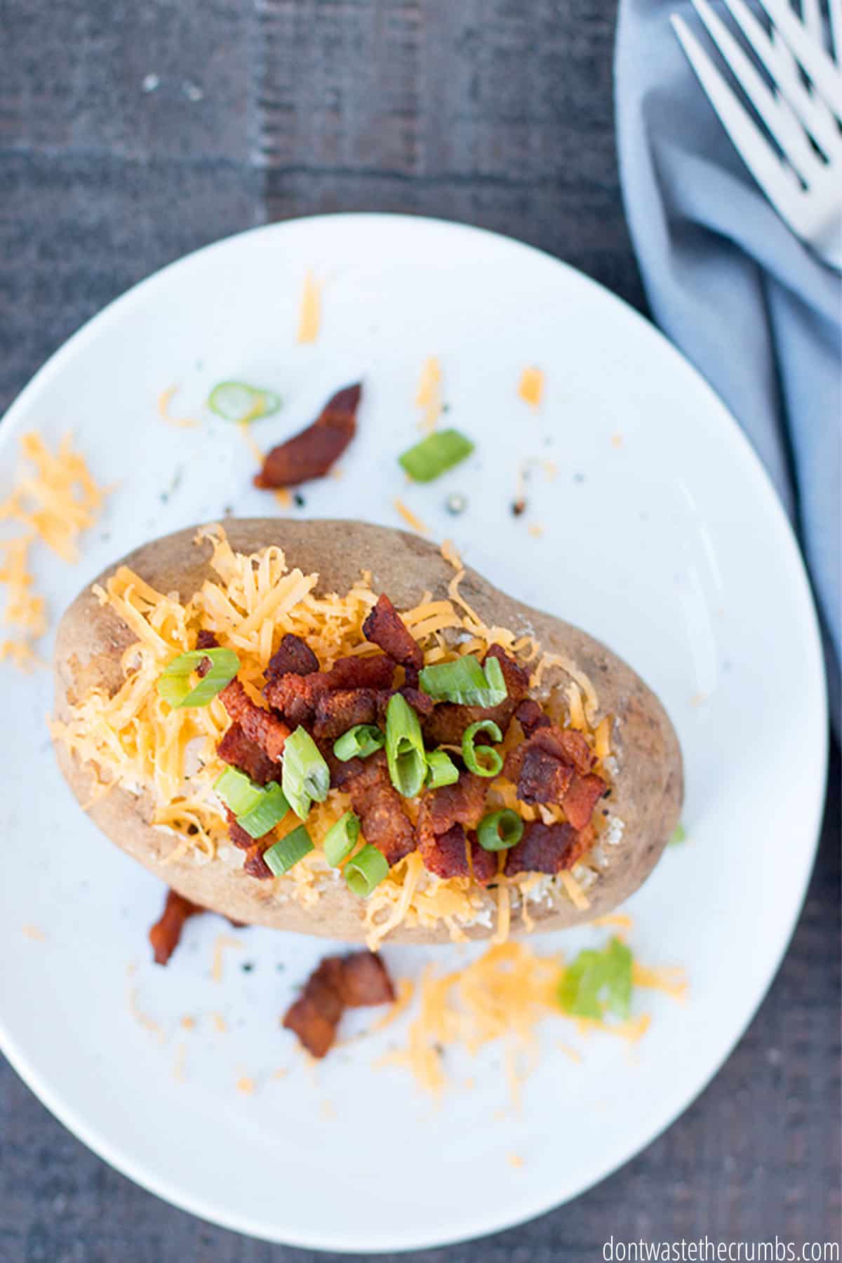 A white plate with a perfectly cooked baked potato, dressed with all of the toppings such as cheese, bacon and green onions.