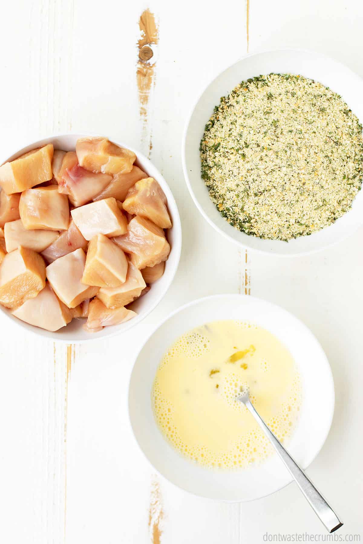 Cubed, 1 inch pieces of raw chicken fill one bowl; a second bowl is filled with whipped eggs, and a third bowl is filled with flour and seasonings.