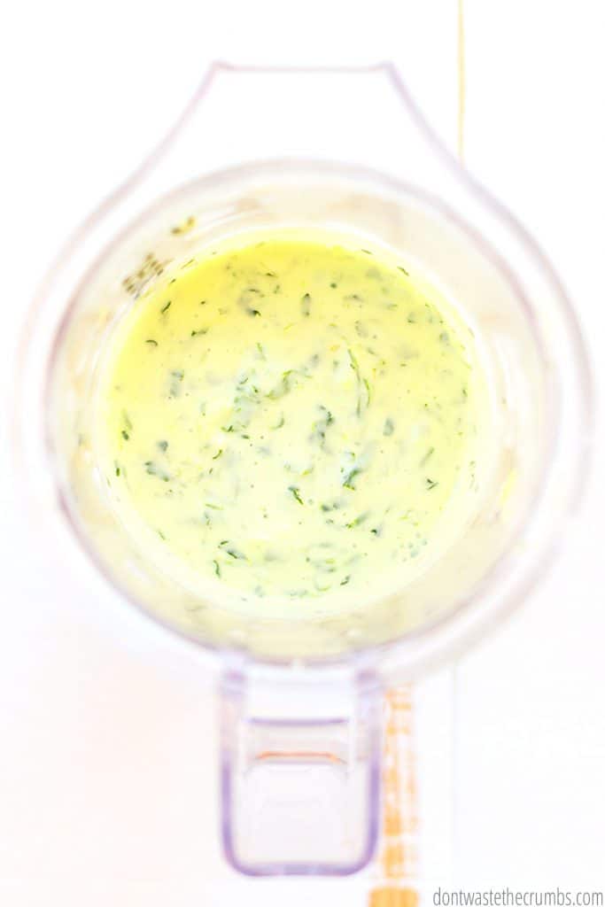 A blender carafe is filled with blended, creamy, cilantro dressing, with pieces of cilantro and green onion visible in the mixture.