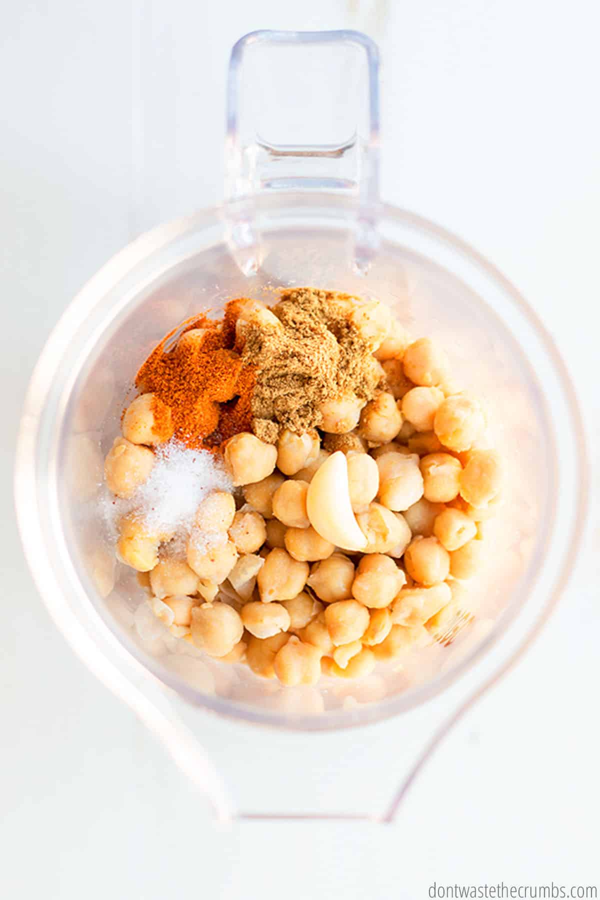 A food processor with the ingredients for homemade hummus (garbanzo beans, garlic, salt, cumin, cayenne pepper & water).