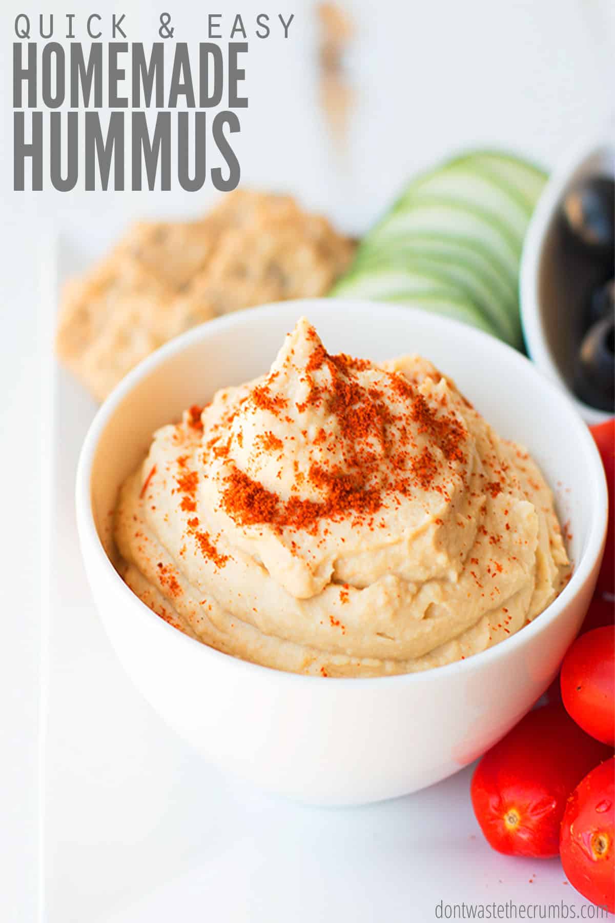 A small bowl of freshly made hummus topped with cayenne pepper. There are crackers, and sliced raw veggies on the side. The text overlay reads "Quick & Easy Homemade Hummus."