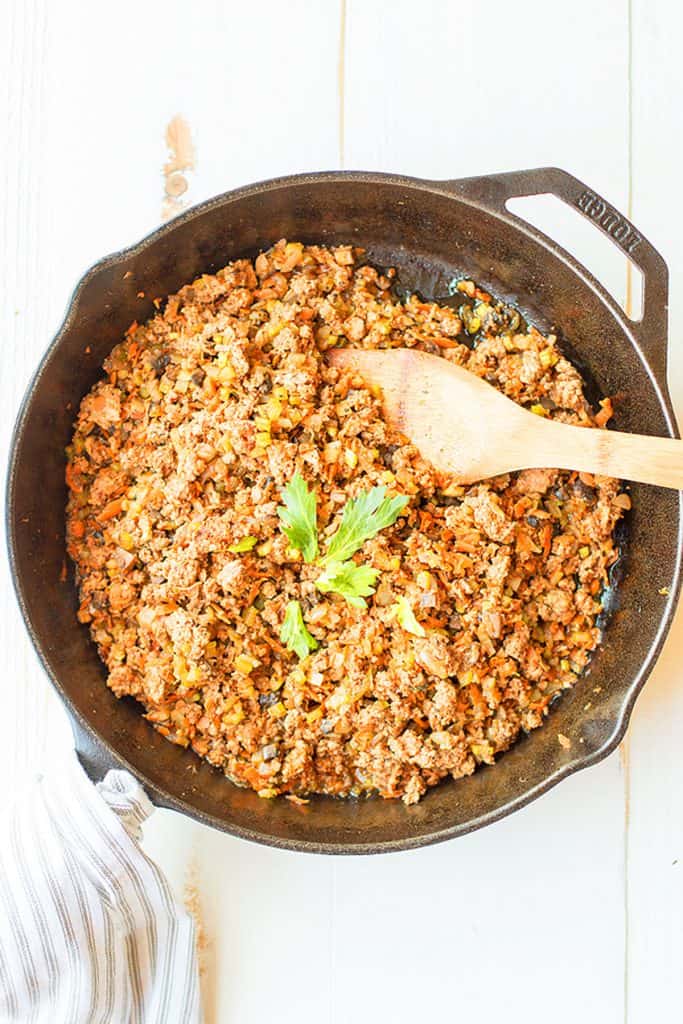 Cooked ground taco meat and vegetables are in a cast iron skillet, along with a wooden serving spoon.