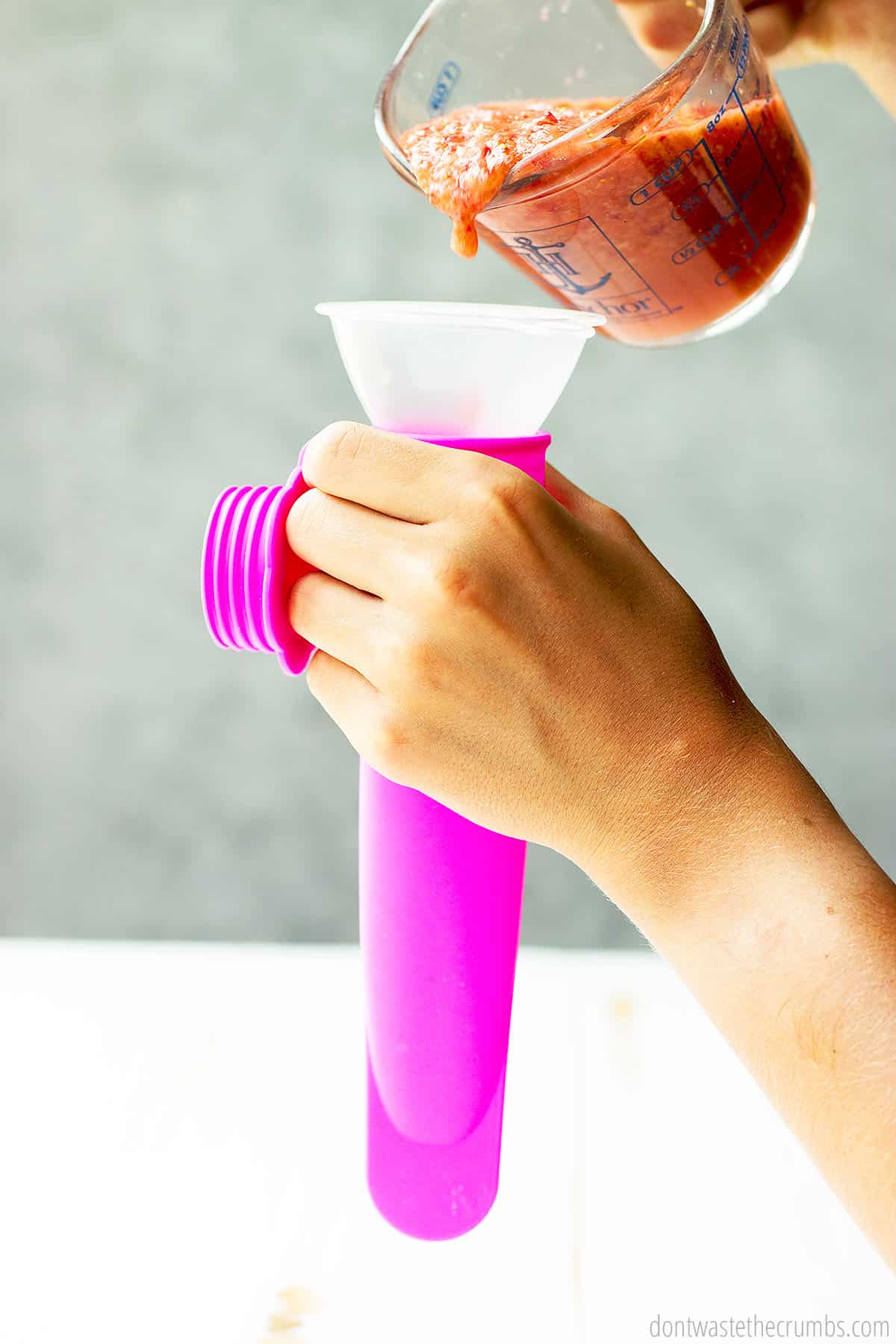One hand holding a reusable ice pop tube with a funnel in the top, the other hand pouring ice pop mix out of a glass measuring cup