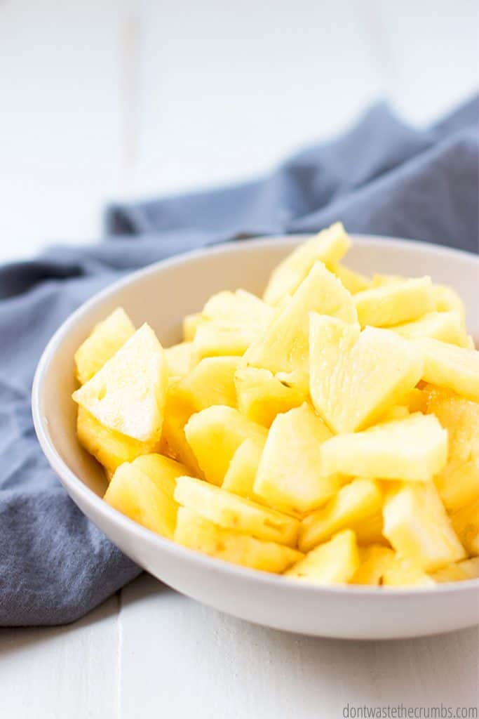 A white bowl filled to the top with freshly sliced pineapple triangles. A blue tea towel sits beside the bowl. 