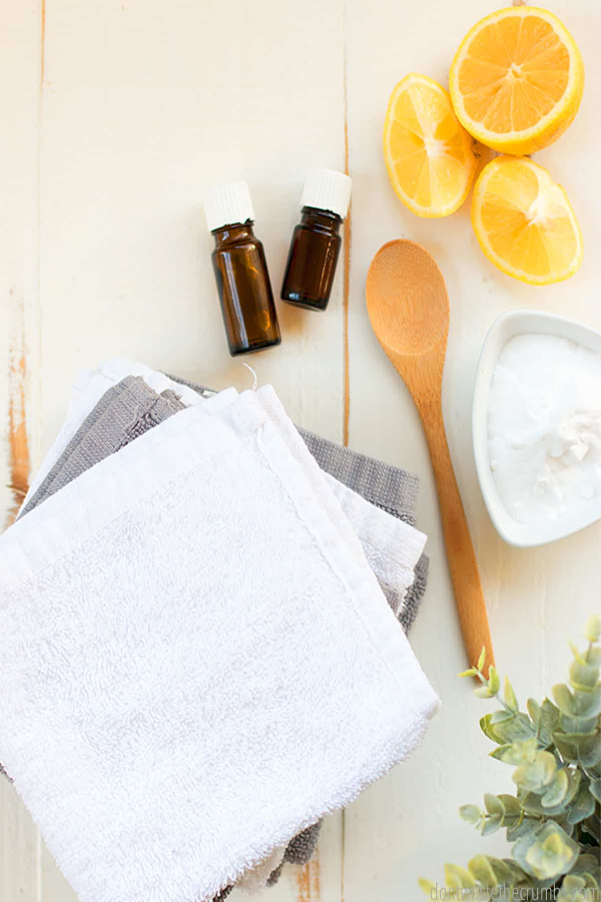 A pile of folded towels lie next to a bowl of baking soda, two bottles of essential oils, and lemon.