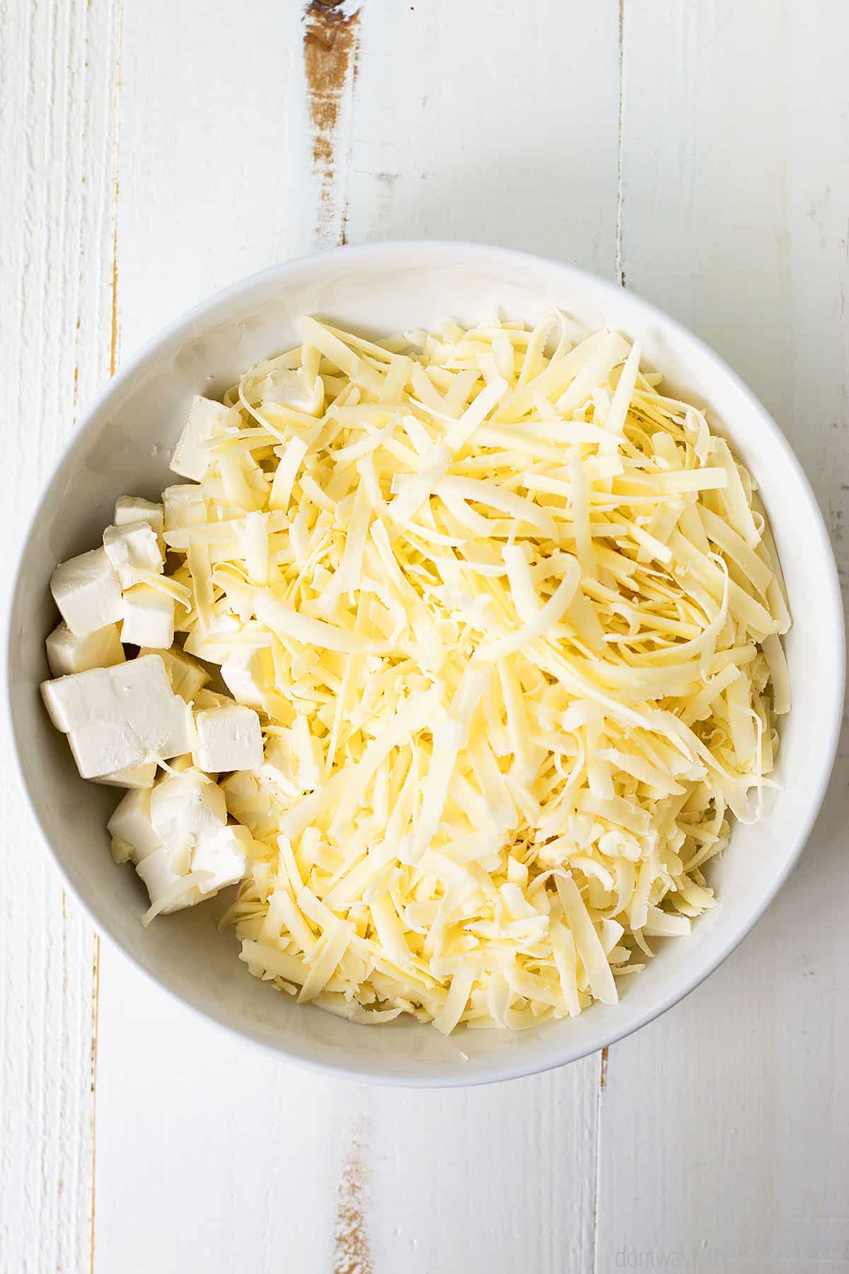 White bowl with shredded cheese and cubed pieces of cream cheese.