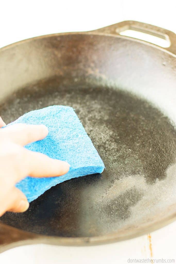 A person is washing a 12" cast iron skillet with a blue sponge in hand.