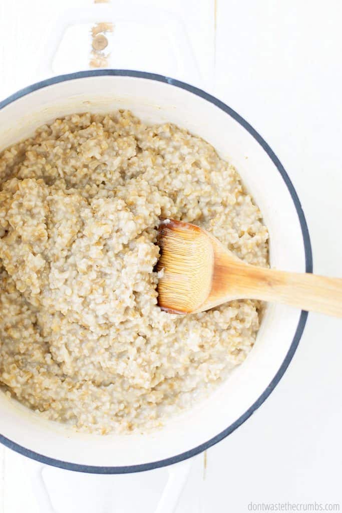 A dutch oven is filled with cooked steel cut oats. A wooden serving spoon is inserted into the oatmeal.