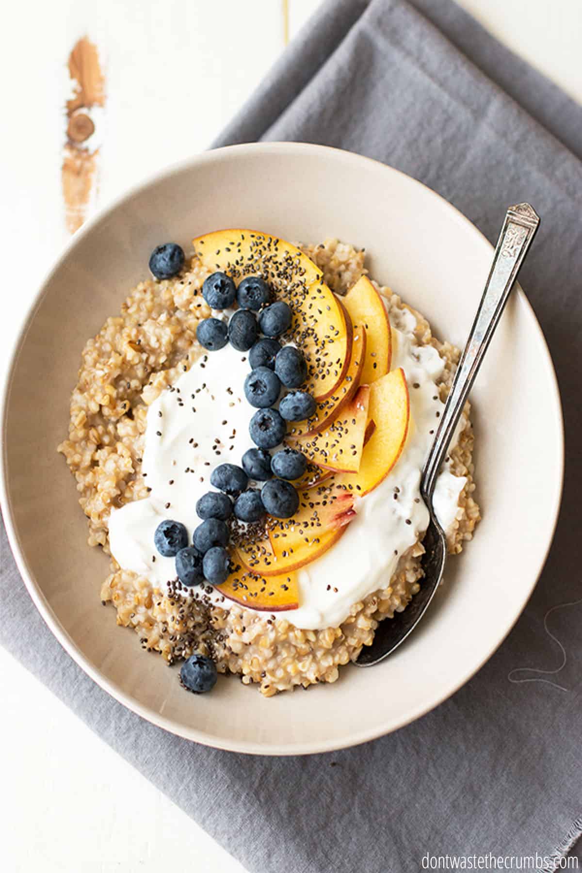 A bowl of cooked steel cut oats is topped generously with yogurt and fresh berries and peaches.