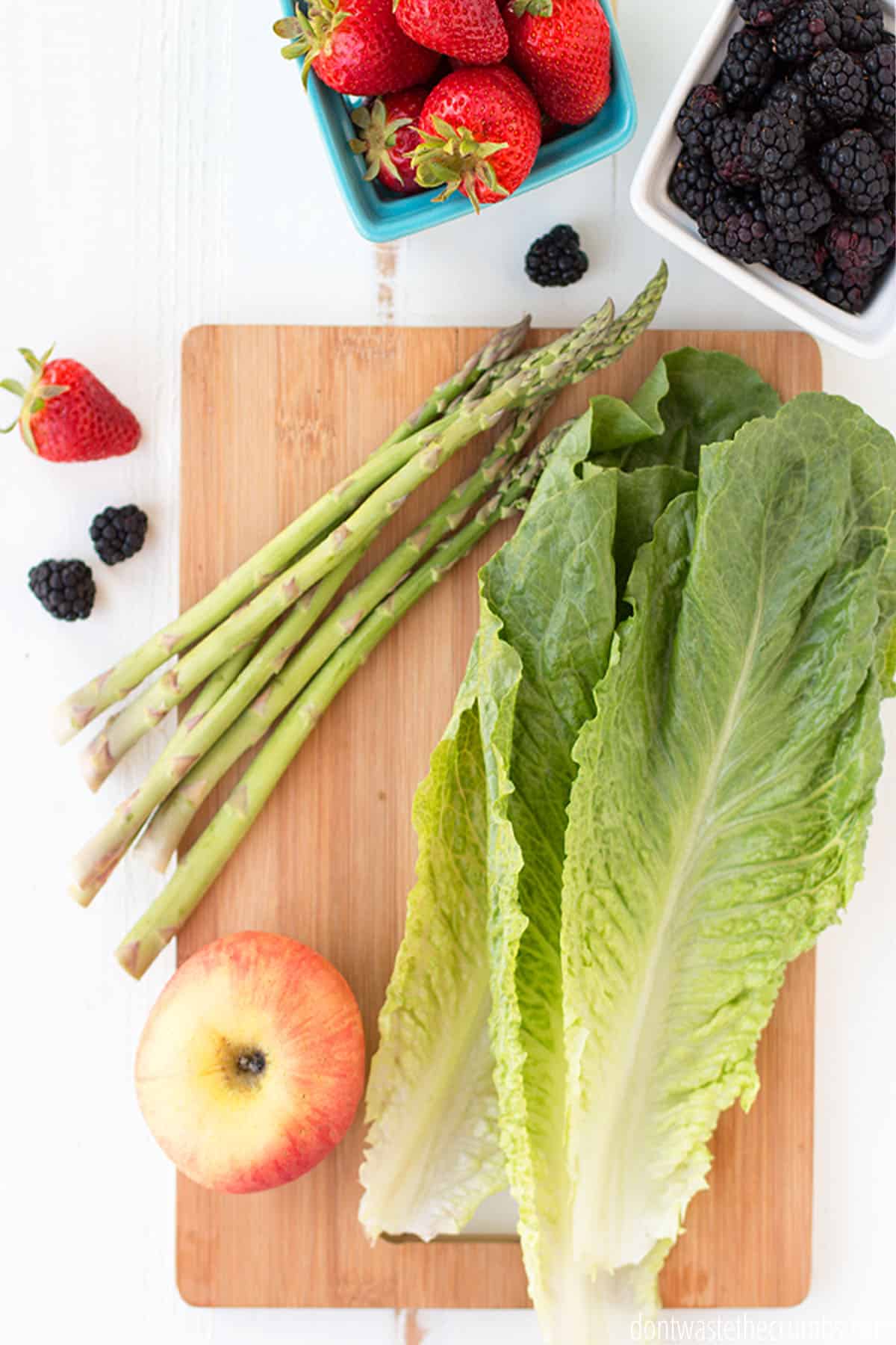 Cutting board with a bunch of asparagus, apple, and lettuce laying on it. On the left there is one strawberry and two blackberries. Above the cutting board there is a bowl of strawberries and a bowl of blackberries.