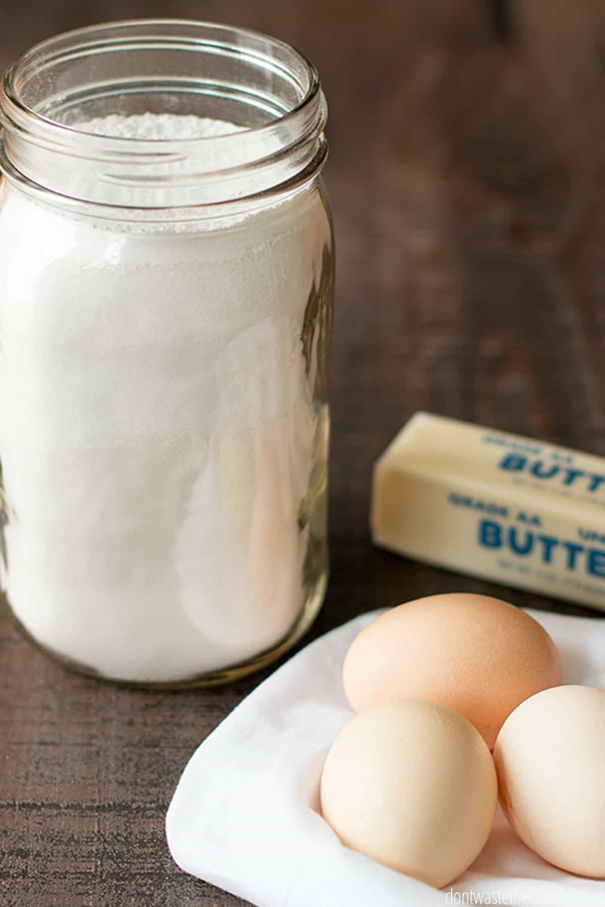 A mason jar of mix, with a stick of butter to the right, and three eggs on a towel