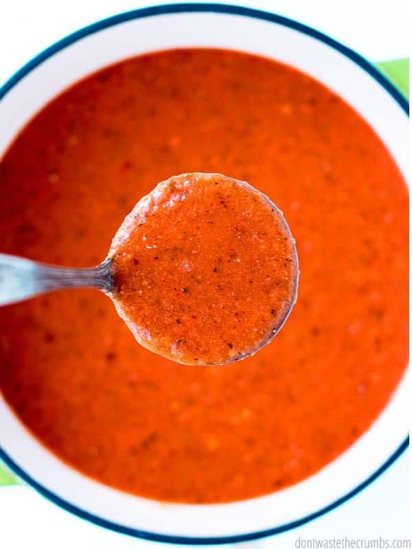 Tomato soup is ladled in a spoon over a bowl filled with deep red broth.