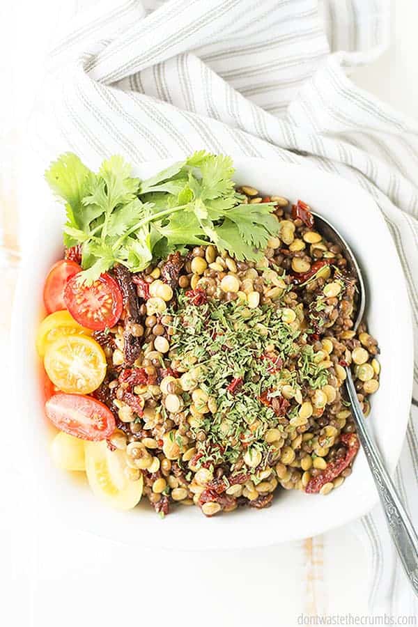 A bowl of marinated lentils is topped with fresh herbs with grape tomatoes and fresh cilantro as garnish.