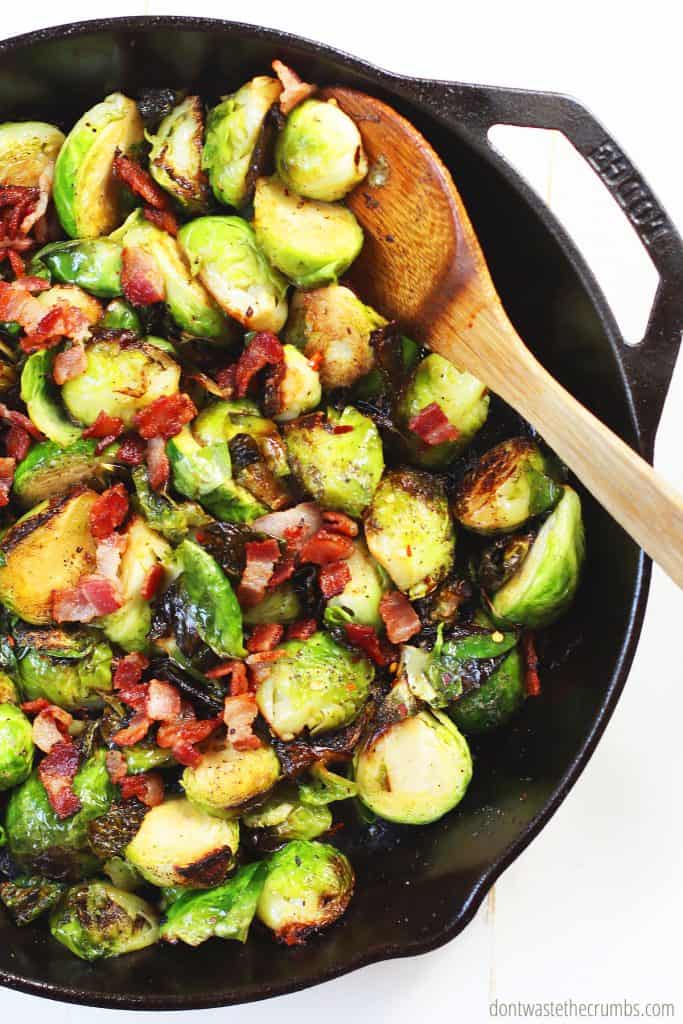 A cast iron skillet with freshly roasted brussels sprouts with bacon and onions. A wooden spoon stirs the Brussels sprouts as they saute.  