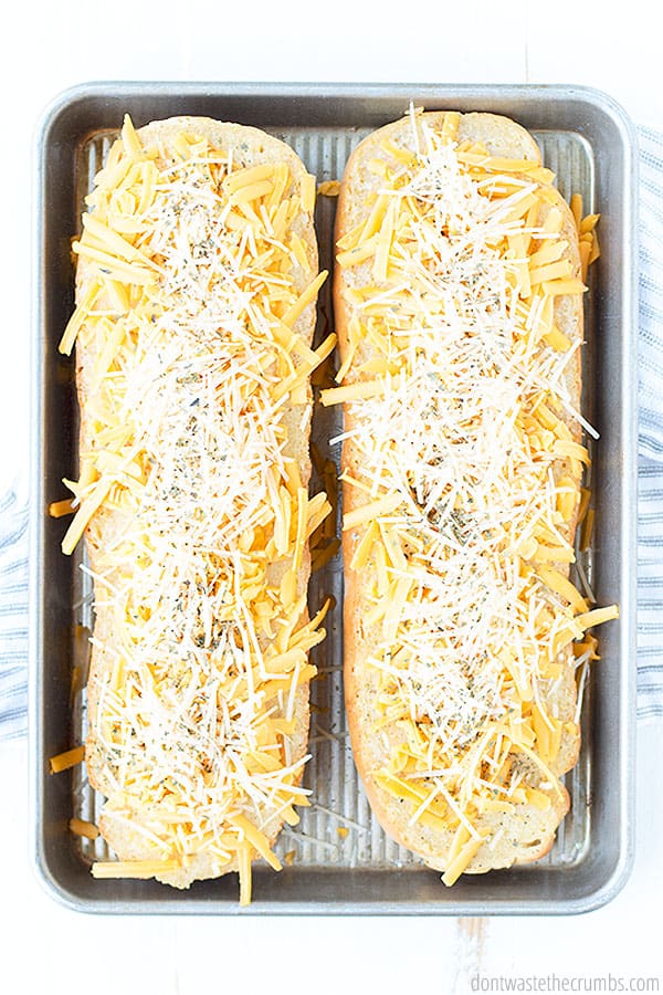 Two different cheeses (cheddar and parmesan) are sprinkled evenly over two halves of garlic butter smeared French bread. They're ready to be baked on a baking sheet in the oven.