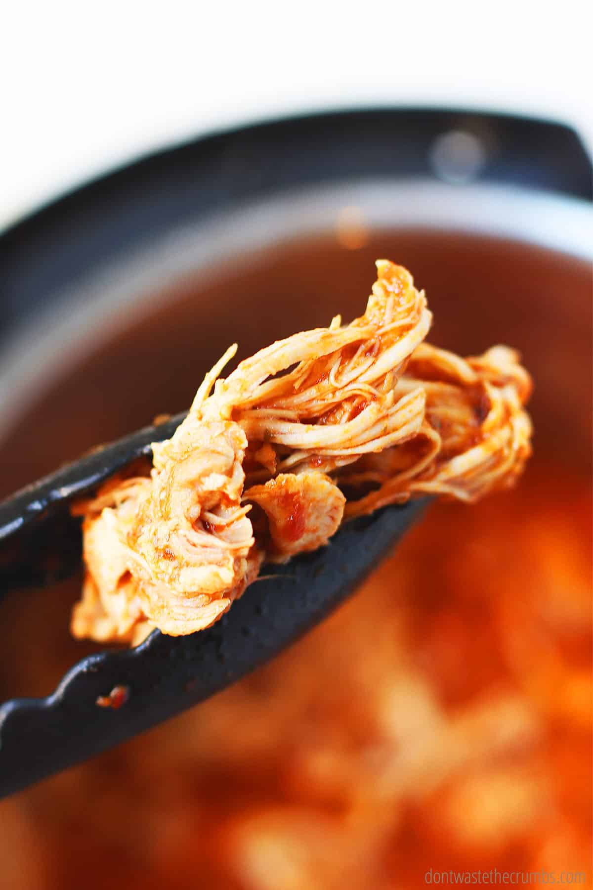 A pair of tongs holding up a piece of cooked chicken tinga with the red adobo sauce in the background.