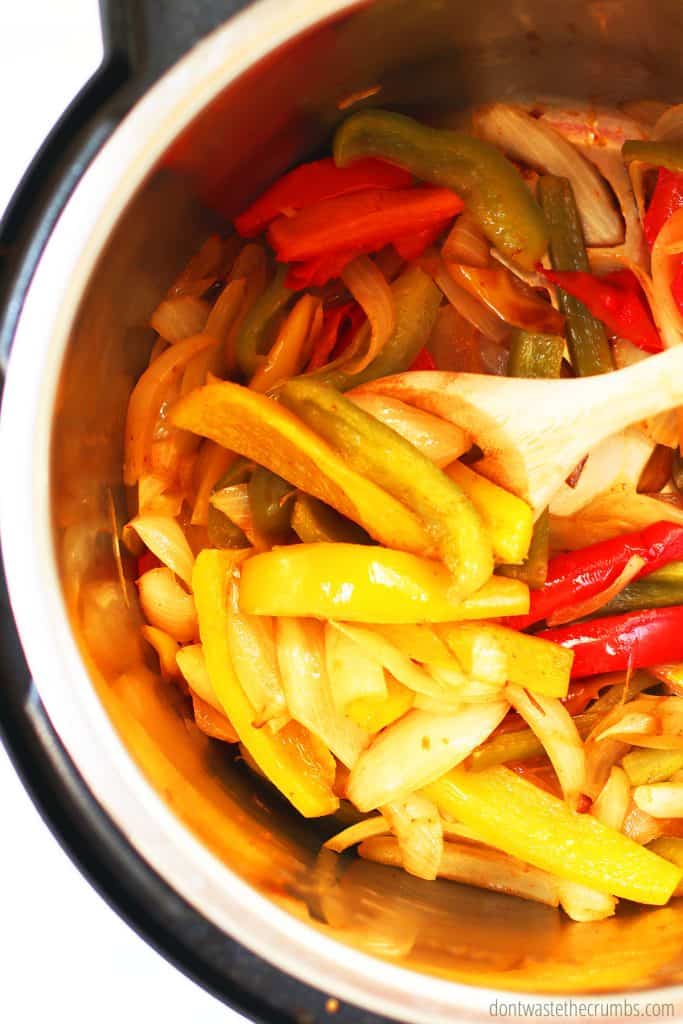 Freshly sauteed sliced bell peppers and sliced onions inside of an instant pot stirred with a slotted wooden spoon. 