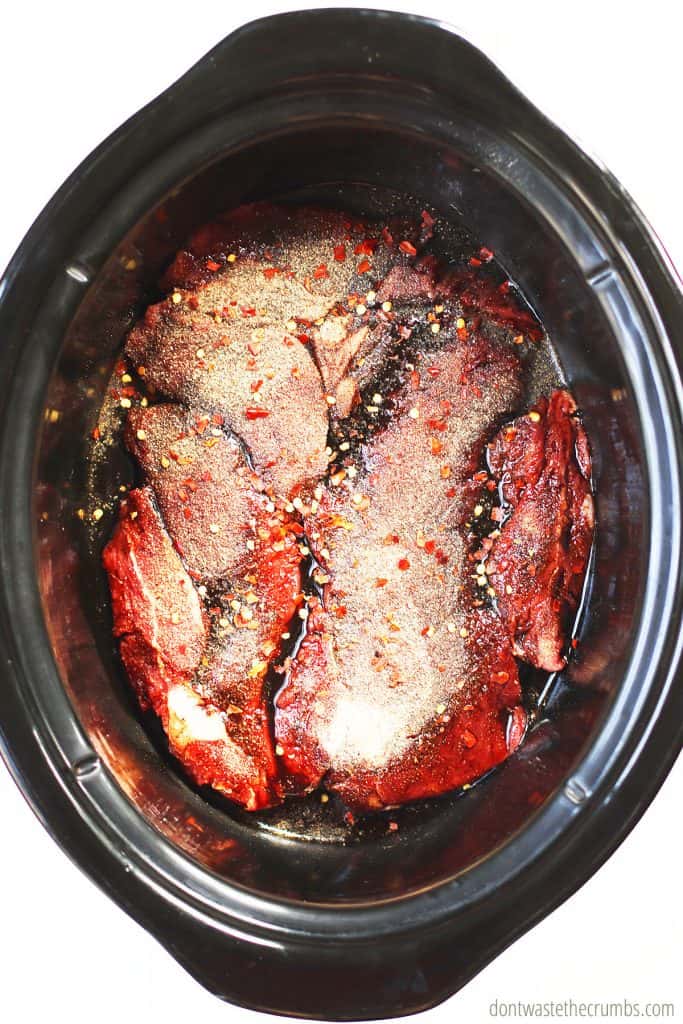 Raw beef roast at the bottom of an oval slow cooker seasoned with balsamic vinegar, red pepper flakes, salt, garlic powder, and black pepper. 
