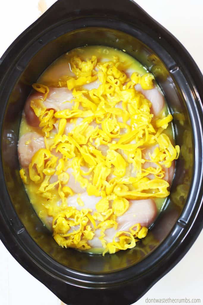 Raw chicken on the bottom of the slow cooker with the cooking liquid poured over, pepperoncini, waiting for the seasonings to be added. 