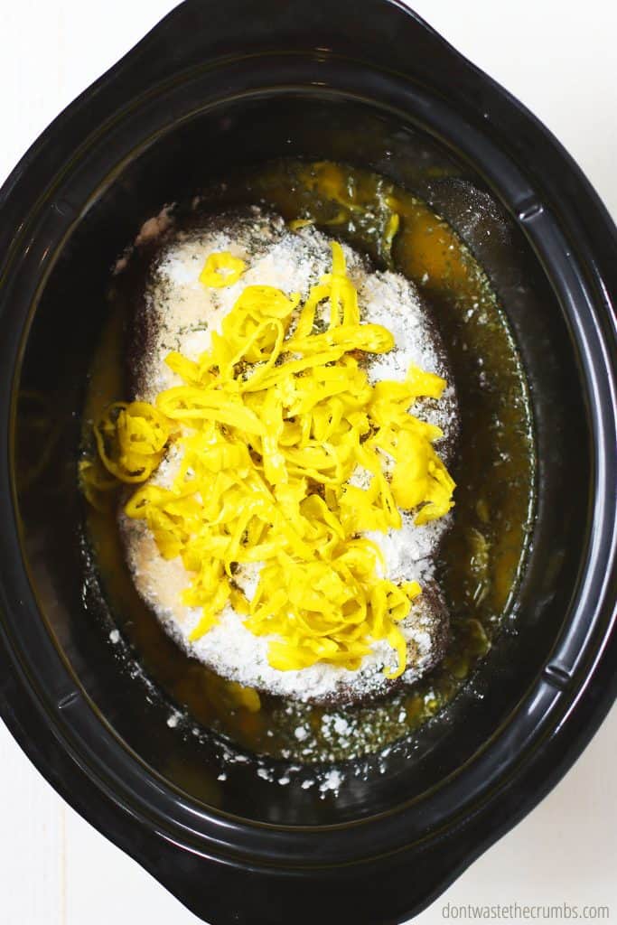 This Mississippi pot roast has sliced pepperoncini peppers sprinkled throughout and dry ingredient mix laying in a slow cooker. 