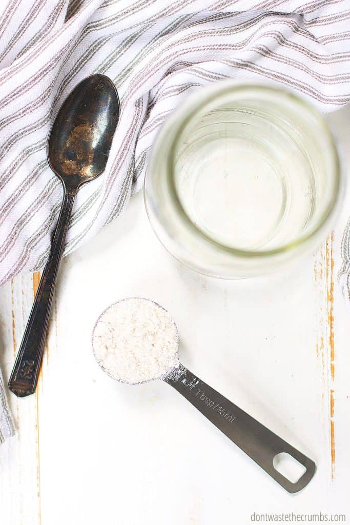 A spoon and tablespoon measuring spoon with brown rice flower and an empty glass jar are on a table. This is what you need to start making gluten-free sourdough starter.