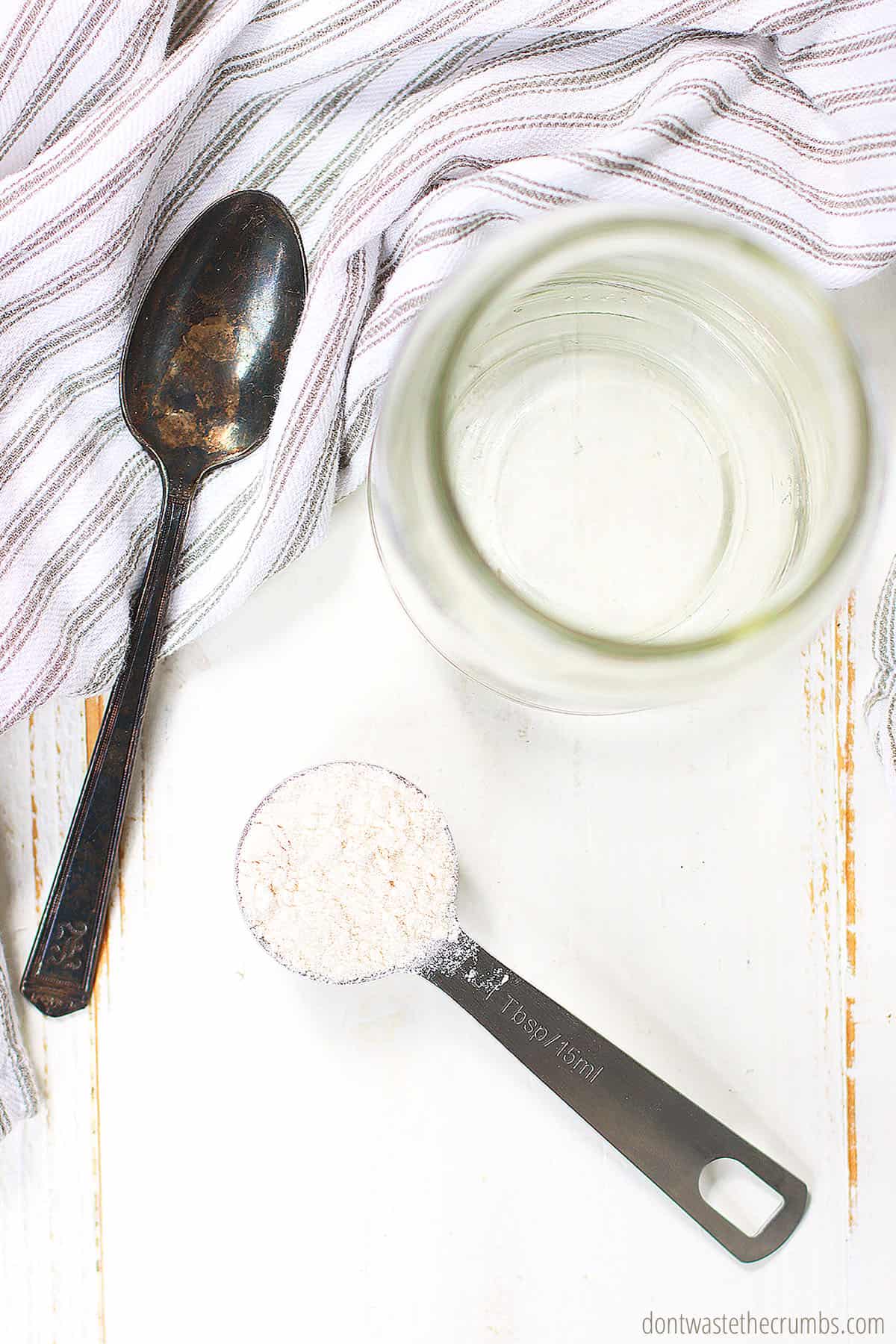 A spoon and tablespoon measuring spoon with brown rice flower and an empty glass jar are on a table. 