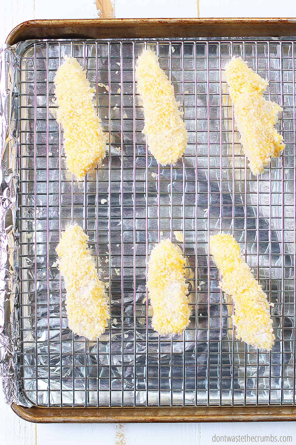 Wire rack over a roasting pan with freshly breaded fish sticks. They are ready to be baked in the oven for 13 minutes.