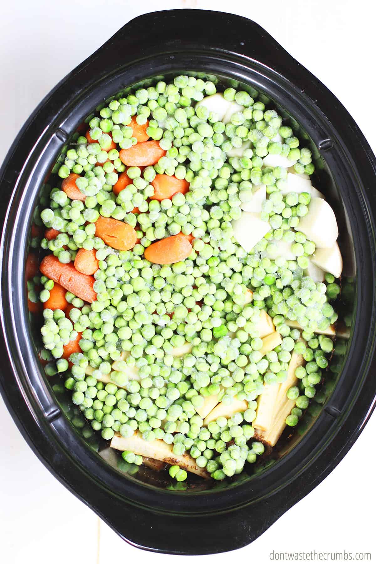 All of the ingredients for this slow cooker beef stew are in the crock pot. Pictured are frozen peas, carrots, parsnips, and turnips.