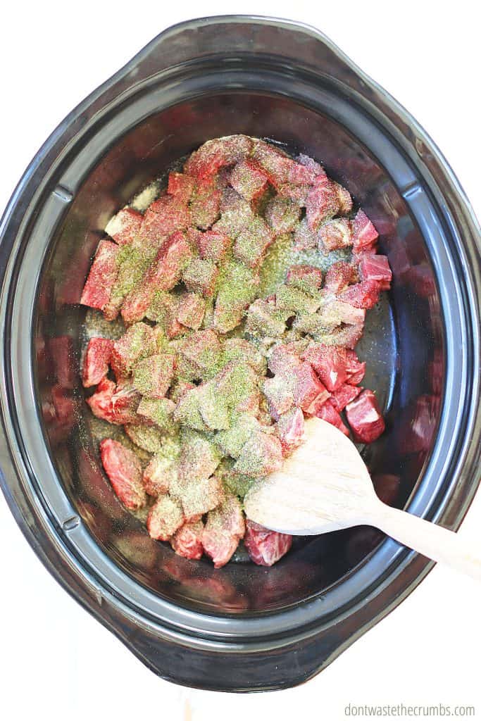 Raw meat that has been cut into chunks are in the slow cooker and there are spices sprinkled through.
