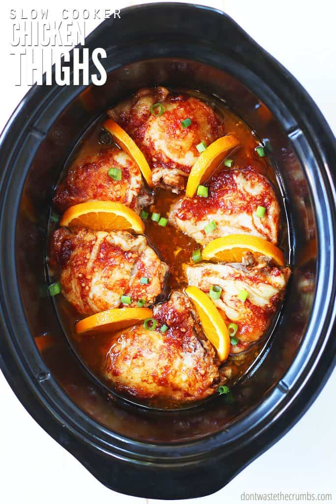 Fully cooked slow cooker chicken thighs in a crock pot garnished with wedges of oranges and sliced green onions. The text overlay reads 'slow cooker chicken thighs.'