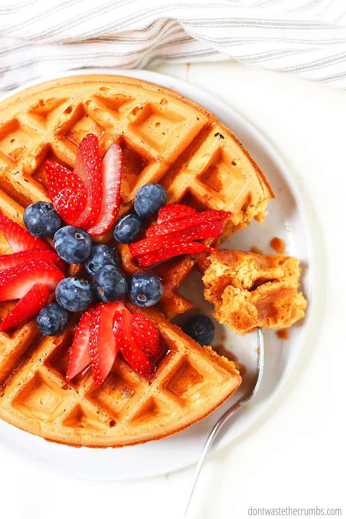 Fresh sourdough waffles with a topping of blueberries and cut strawberries on top. There is a bite taken from the waffles. This delicious breakfast is on a white plate.