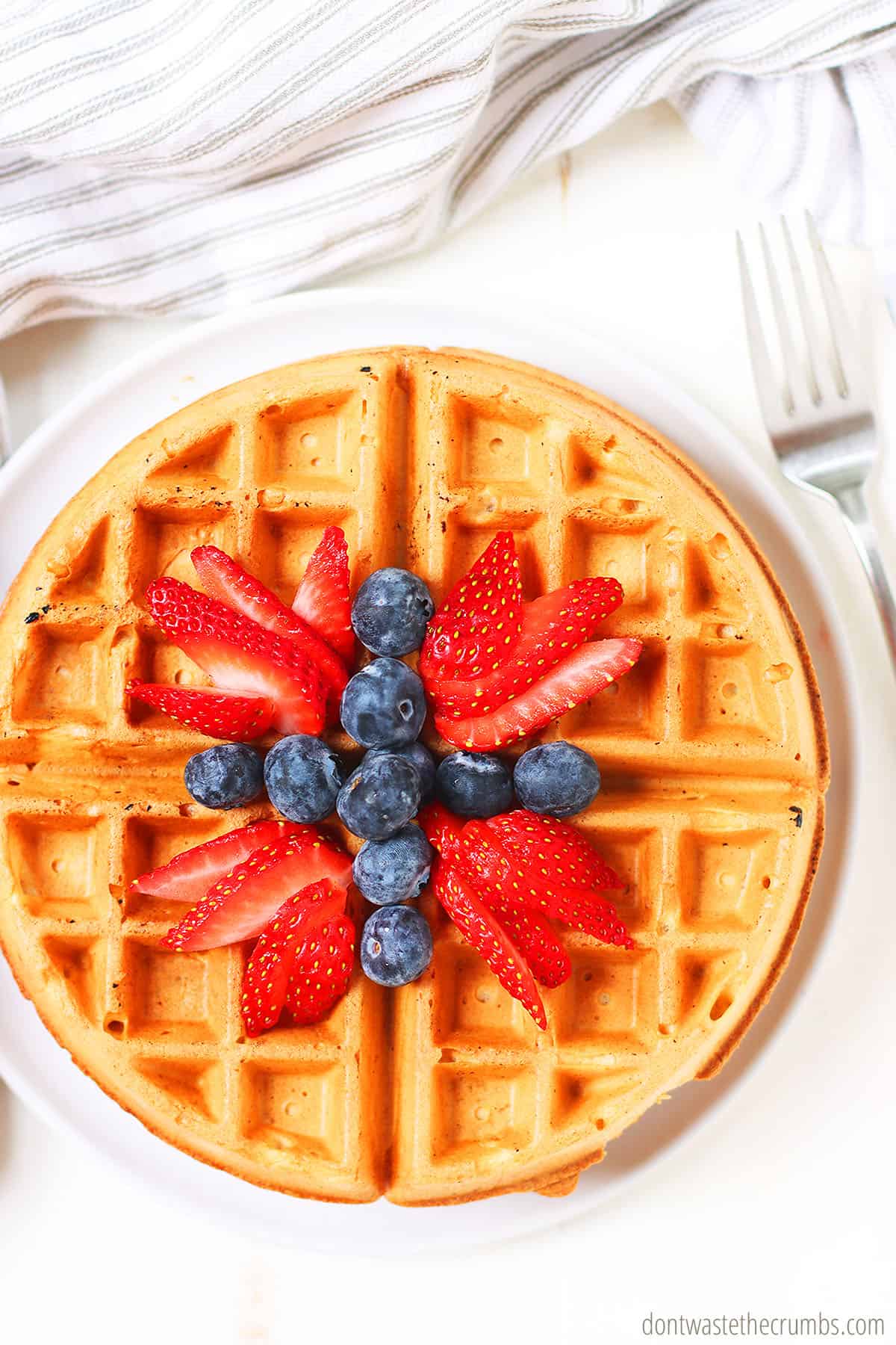 Fresh sourdough waffles with a topping of blueberries and cut strawberries on top. This delicious breakfast is on a white plate.