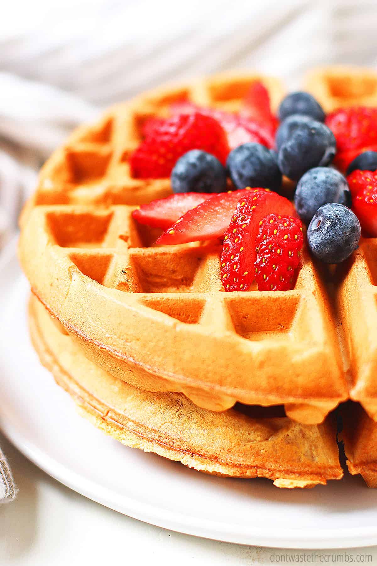 Up close view of fresh sourdough waffles with a topping of blueberries and cut strawberries on top. This delicious breakfast is on a white plate.