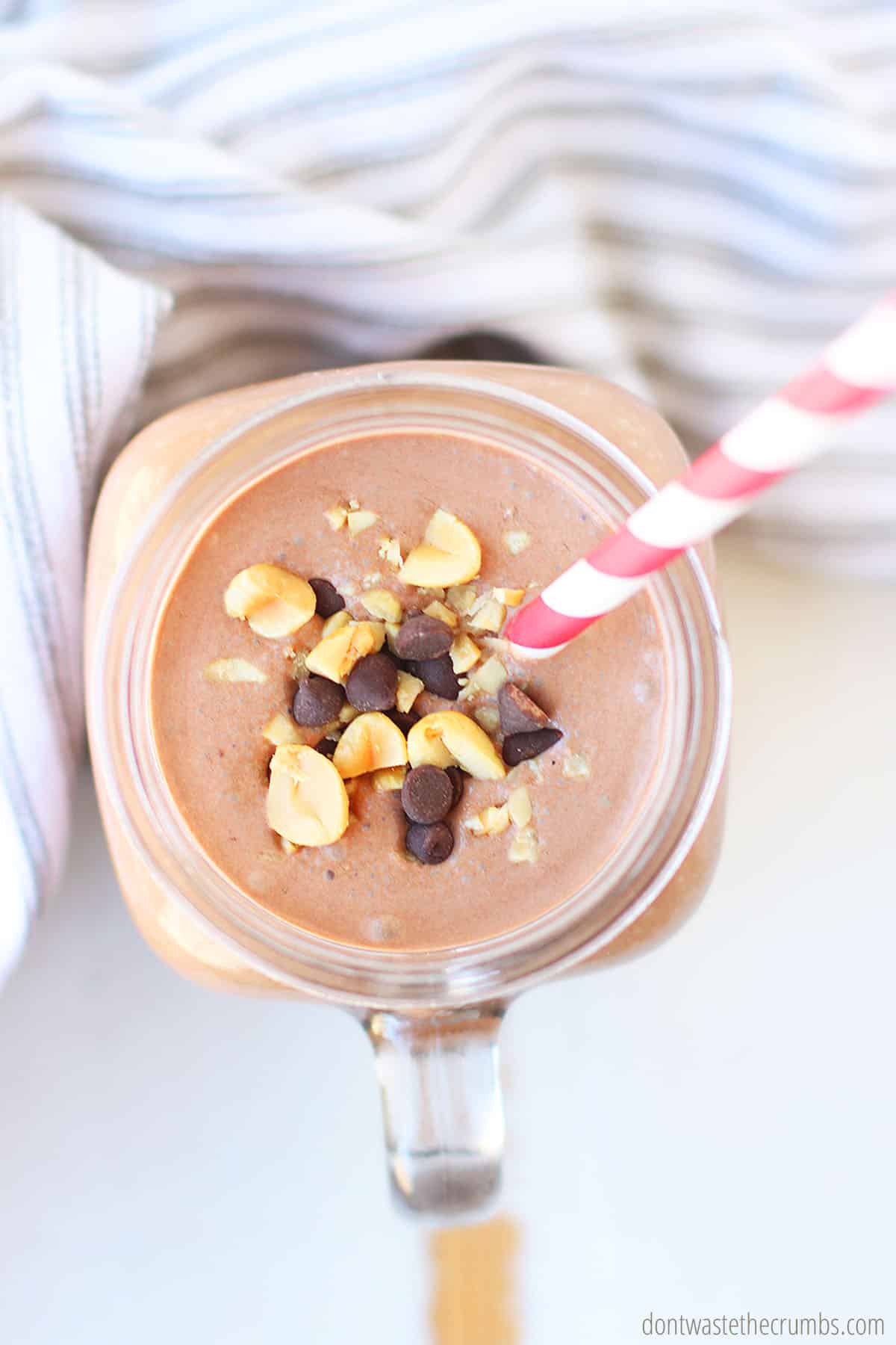 Looking straight down at the top of a peanut butter chocolate smoothie in a glass jar with a red and white striped straw. Garnished with chopped peanuts and chocolate chips.