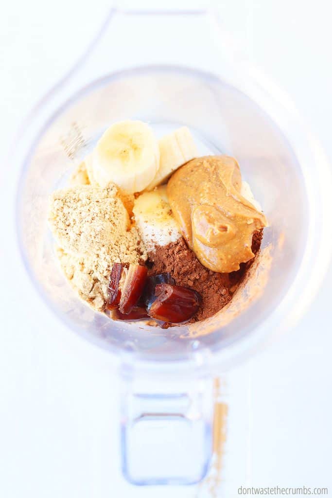 Sliced bananas, peanut butter, peanut butter powder, cocoa powder, dates, and milk inside of a blender ready to be blended into a homemade chocolate banana smoothie. 