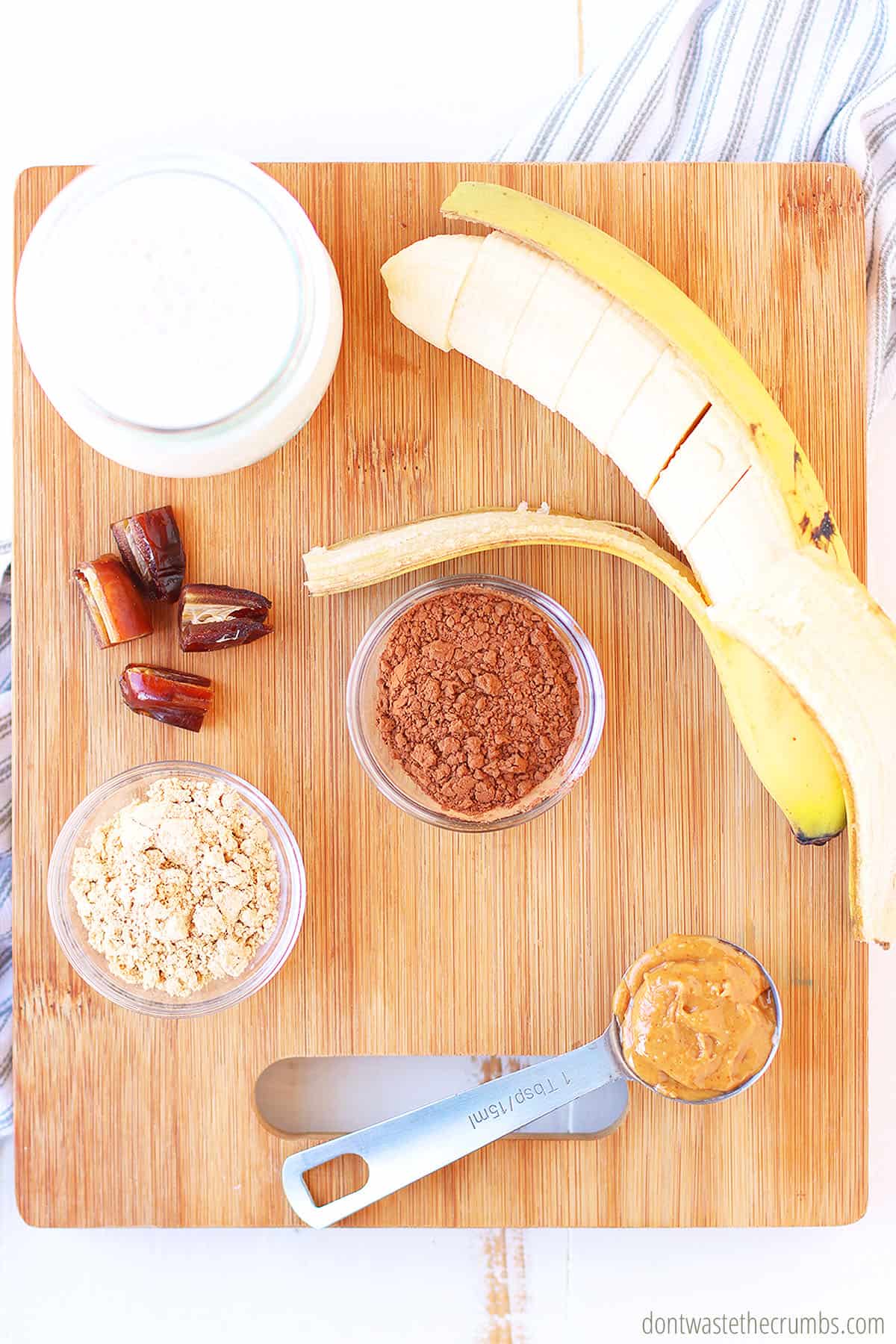 A wooden cutting board with a small cup of milk, sliced banana, sliced dates, peanut butter, cocoa powder, and peanut butter powder.