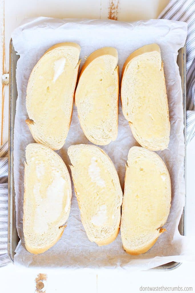 6 slices of sourdough bread on a baking pan with parchment paper. Butter is on top of the slices and ready to be toasted and become crostini!