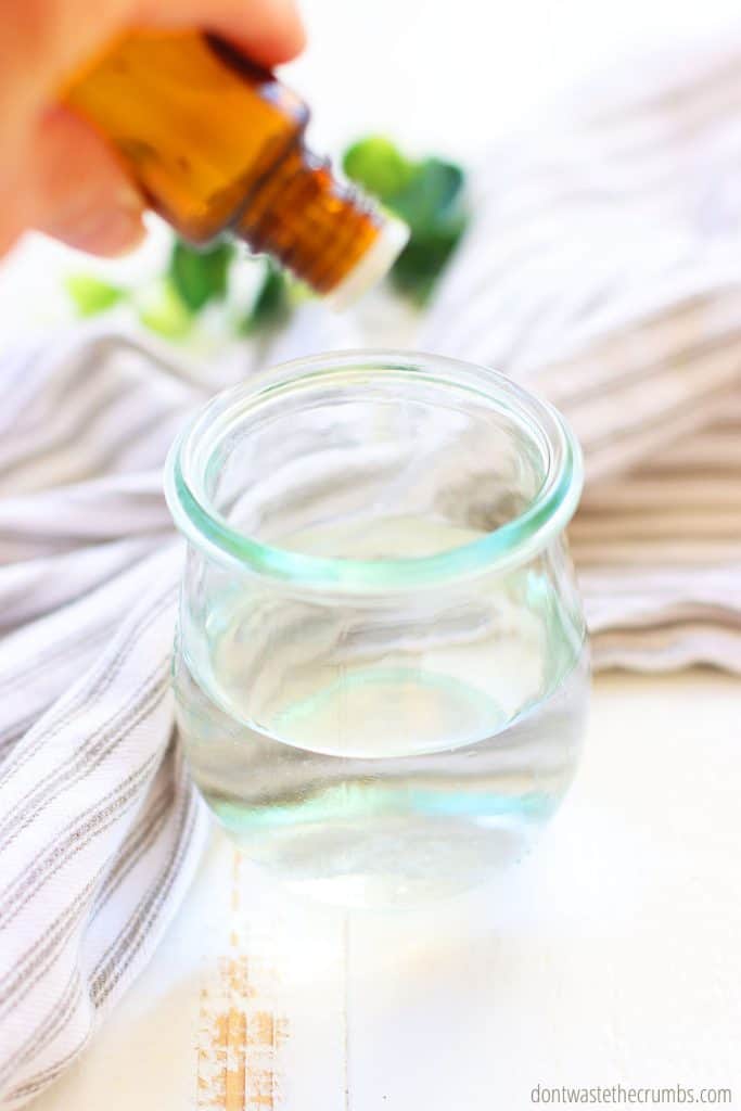 Glass jar with distilled water and a hand tilting an essential oil bottle while drops go into the water. 