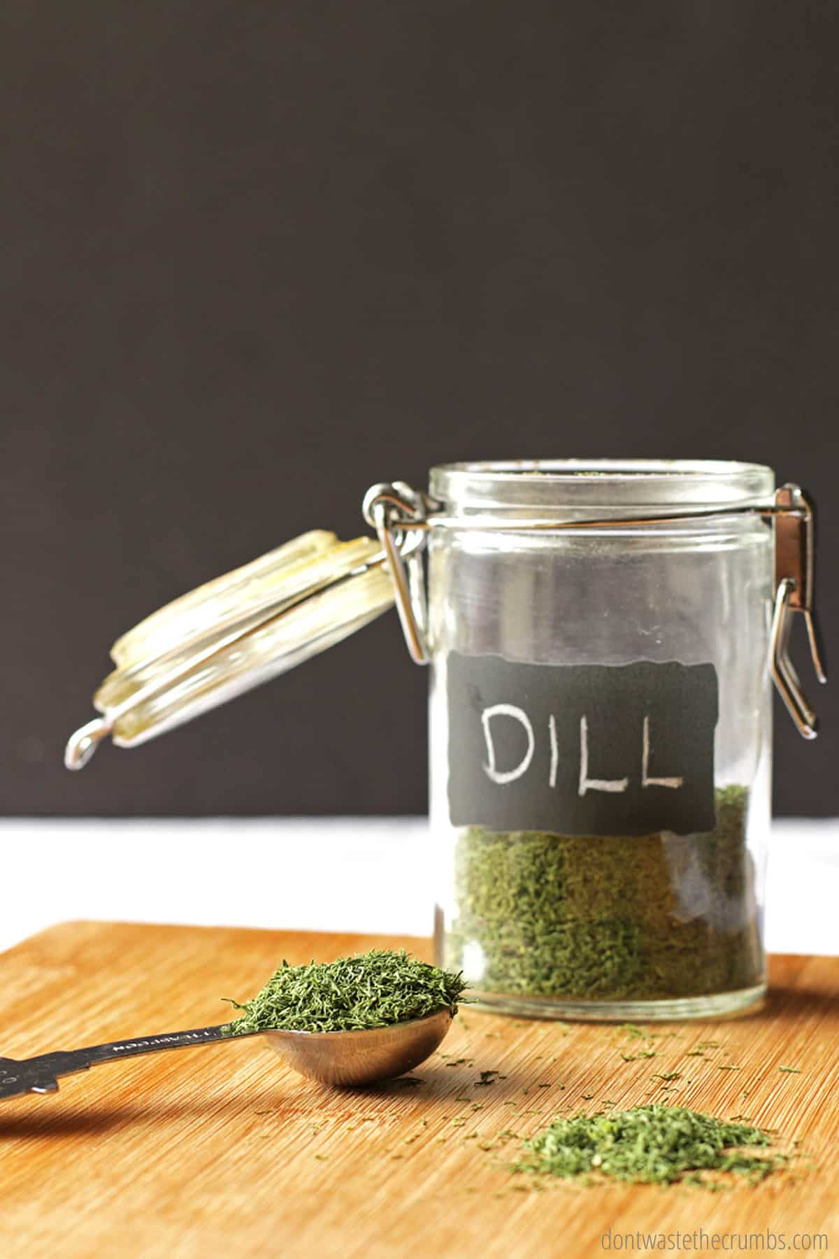 An open jar of dried dill, with a handwritten chalkboard label, sits on a wooden cutting board, with a measuring spoon full of dill to the left and a small pile of dried dill in front