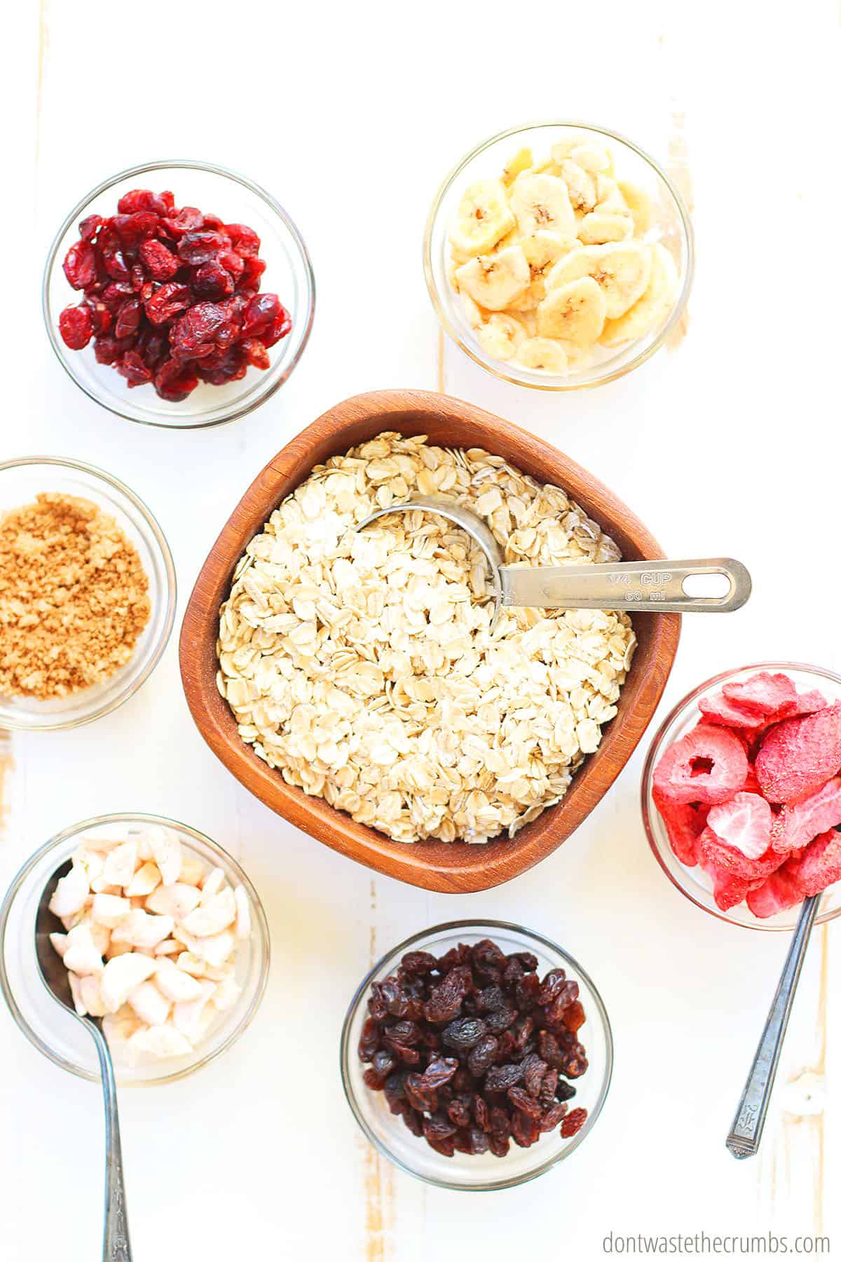 A bowl of oats in the middle and a scoop. Six small bowls are surrounded by the middle bowl. They are mix ins such as, dried strawberries raisins, pecans, dried bananas, brown sugar, and cranberries.