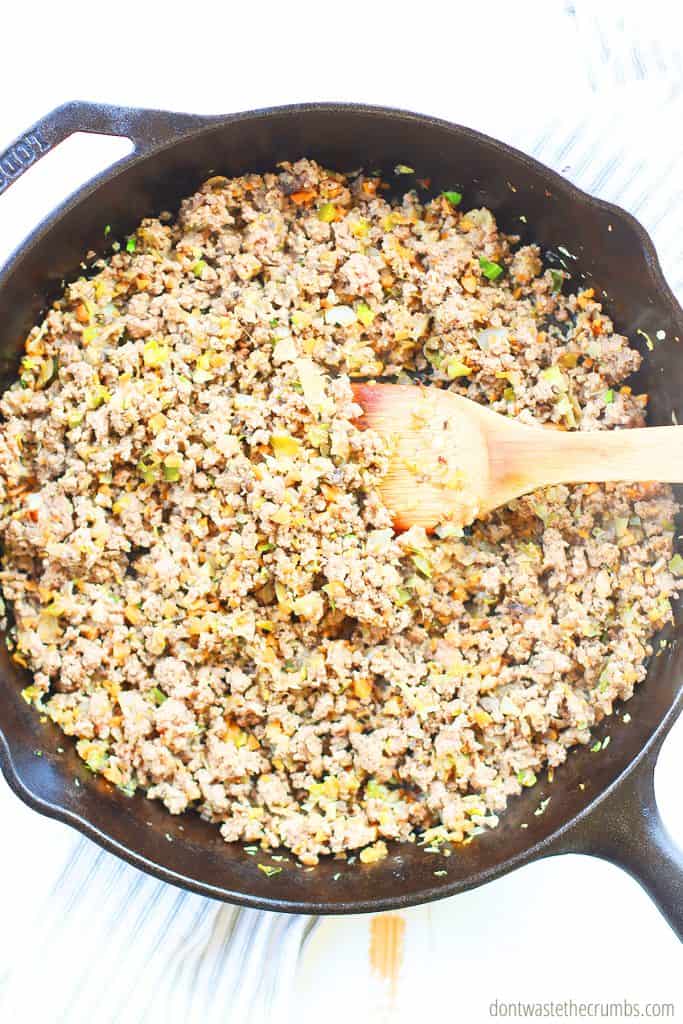 Ground beef mixed with meat fillers that is cooked in a cast iron skillet with a wood spoon stirring.