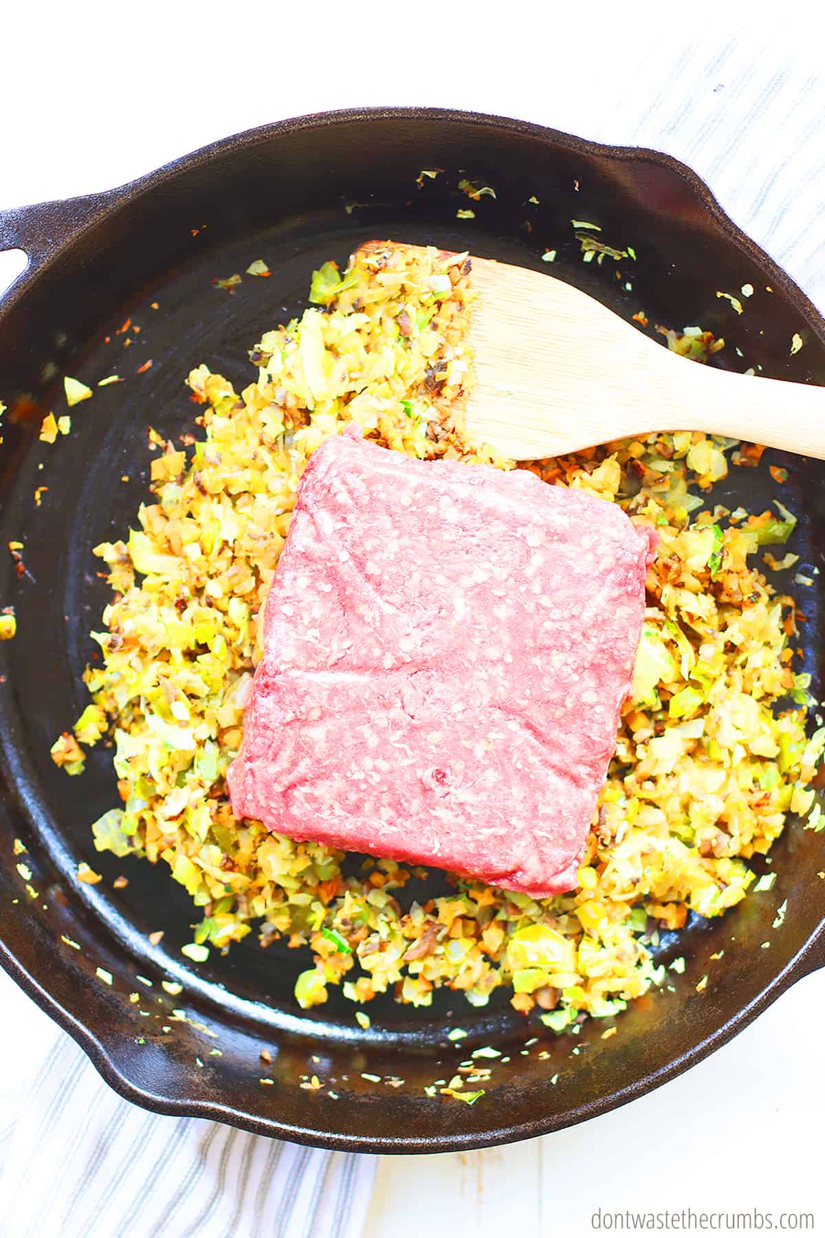 Raw meat on top of the meat filler mixture in a cast iron skillet with a wood spoon stirring.