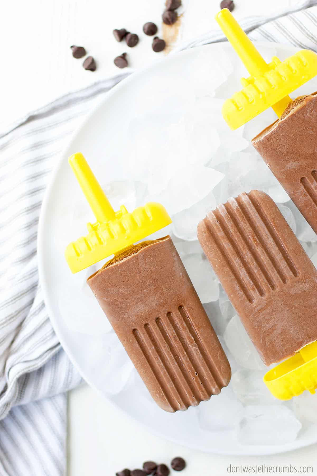 Homemade fudge pops lying in a bowl of ice, on top of a dish towel, with some chocolate chips scattered nearby