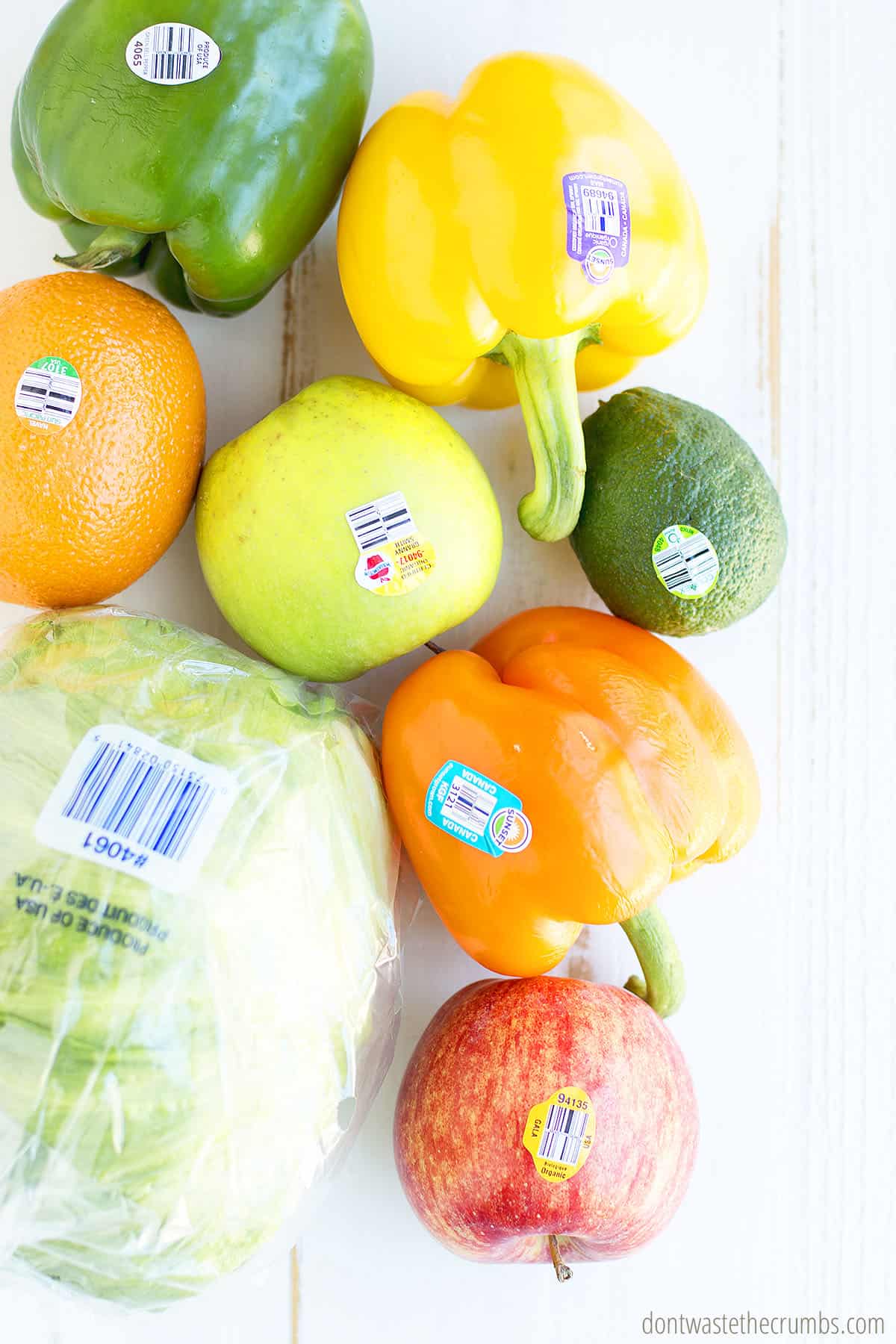 An orange, green pepper, yellow bell pepper, avocado, green apple, orange bell pepper, and lettuce with PLU stickers on them on a table.