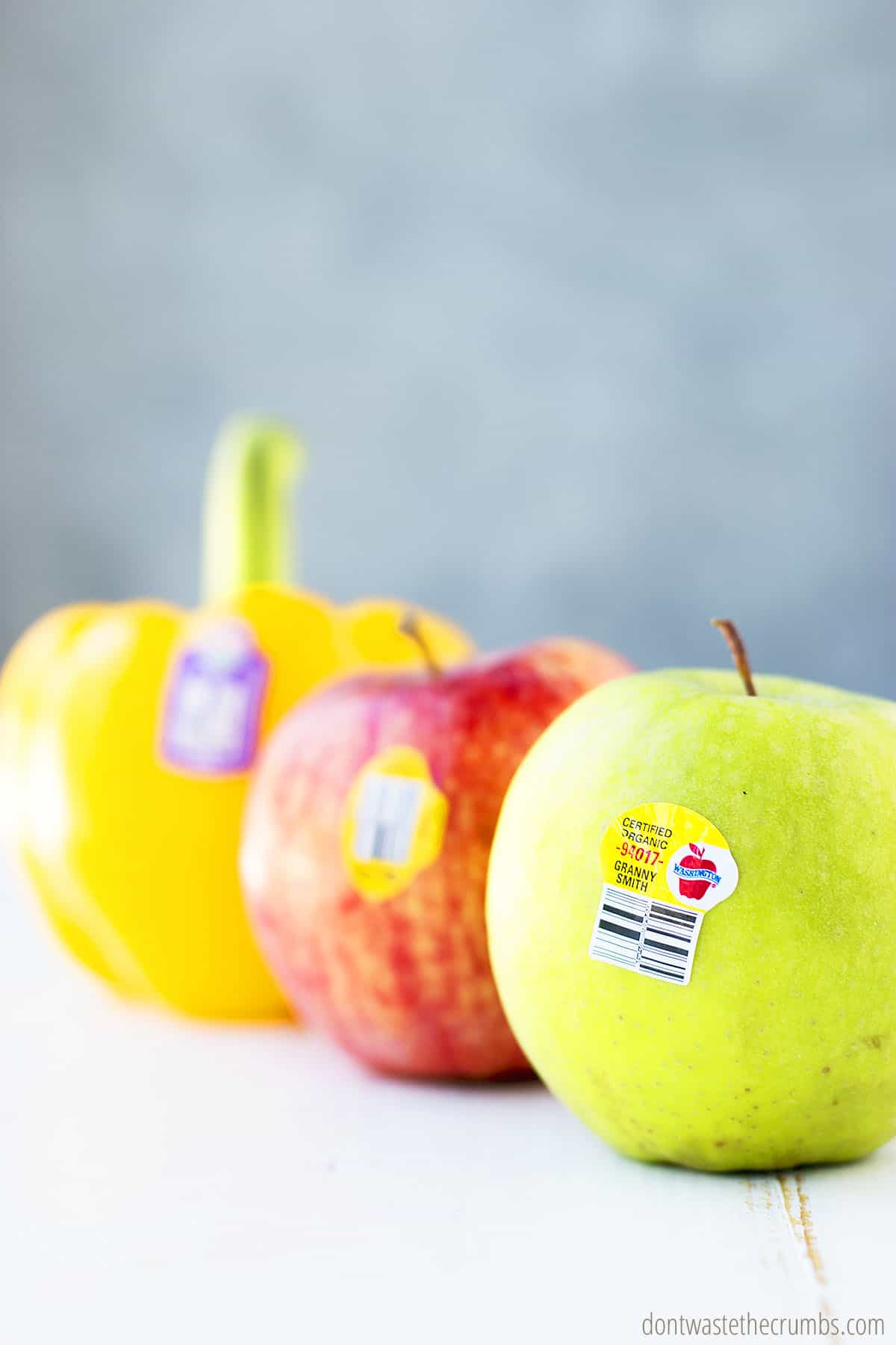 Granny smith apple, red apple, and yellow bell pepper and on a table with PLU stickers.