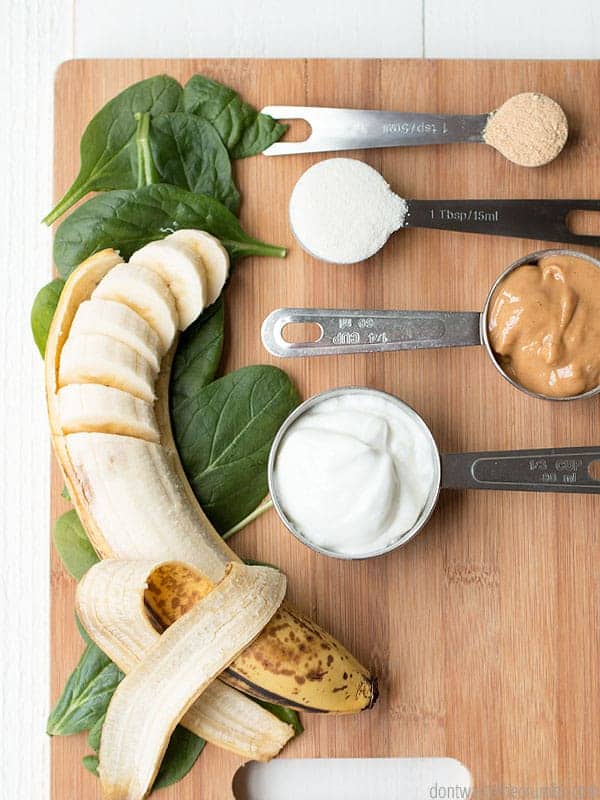 Ingredients for green maca smoothing in three different measuring spoons. Sliced banana on top of spinach leaves are next to the measuring spoons. Everything is on top of a wooden cutting board.