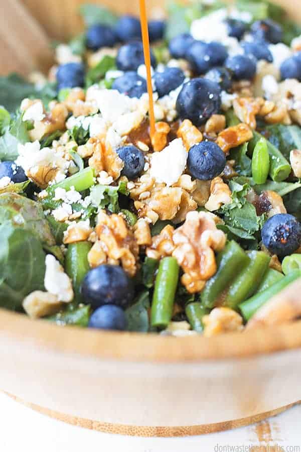 Kale salad with blueberries, walnuts, and feta in a big wooden salad bowl. Salad dressing is being drizzled onto the salad.